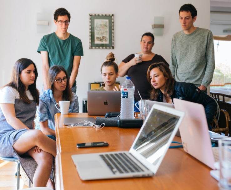 A group of eight young adults crowd around a laptop.