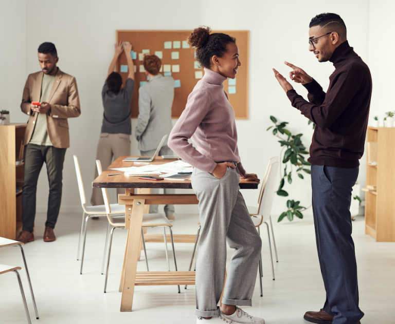 A group of 5 people collaborate in a room that’s set up with tables, chairs, a corkboard, sticky notes, paper, and laptops.