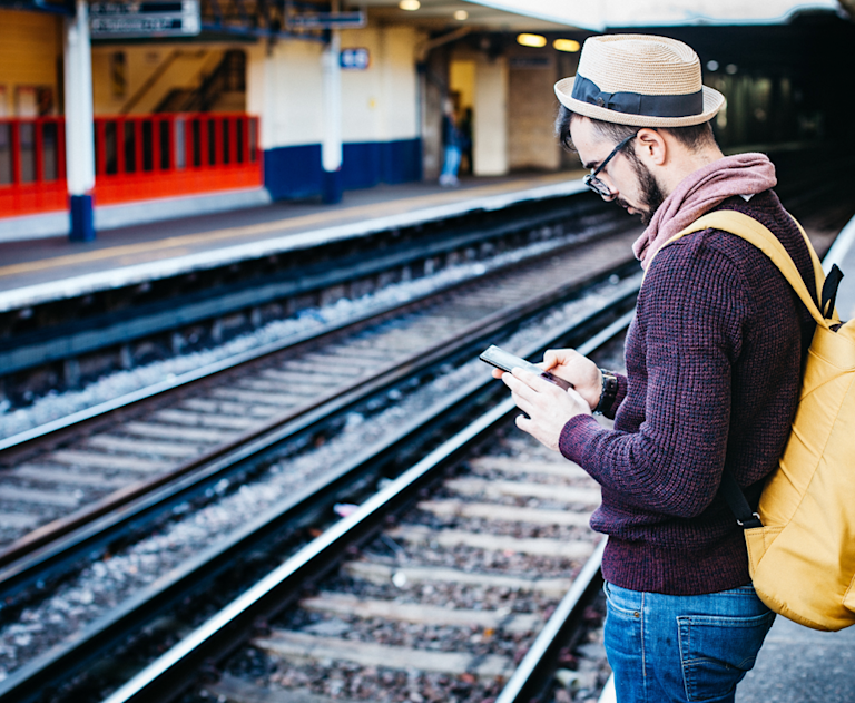 Man stands on a train station platform and looks down at a mobile phone.