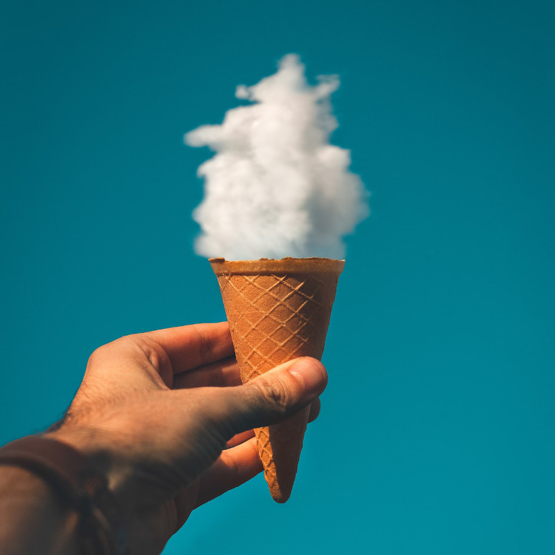 Hand holding an empty ice cream cone creates an optical illusion using a tiny cloud in the sky that looks like ice cream filling the cone.