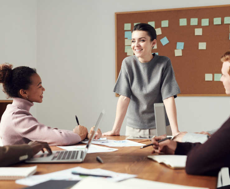 Four people meet in a room in front of a cork board covered in notes. They’re smiling at each other as they sit or stand at a table with laptops, notepads, and pens.