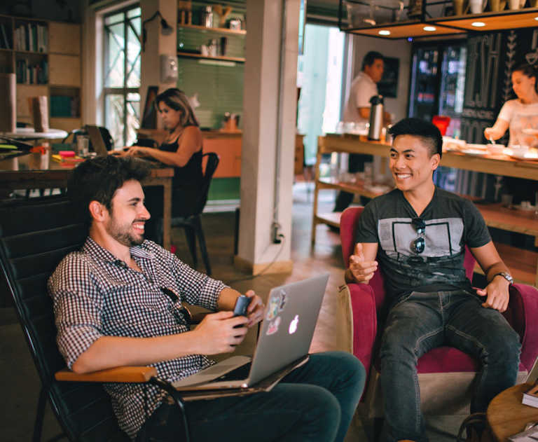 Two men chat while sitting on chairs inside a hip-looking startup office environment. People in the background are working on laptops and serving themselves food.