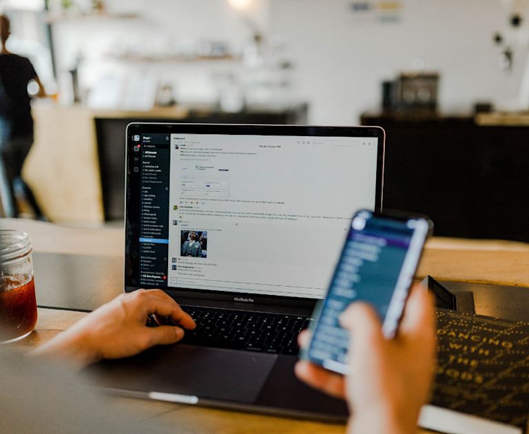 A mobile employee uses Slack on a laptop, while working in a cafe. They’re holding their phone while typing a message on Slack.