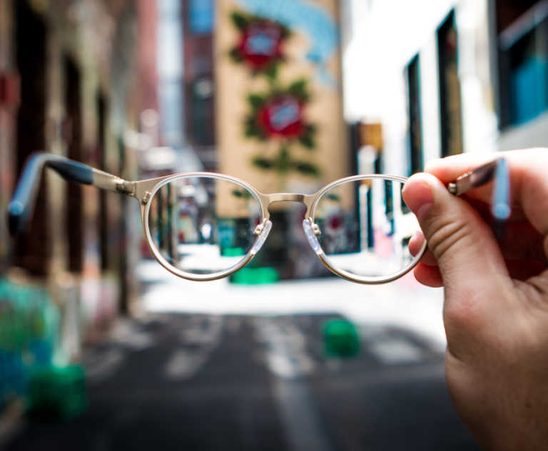 A person holds up a pair of glasses while in an urban setting, with just their hand visible in the image.