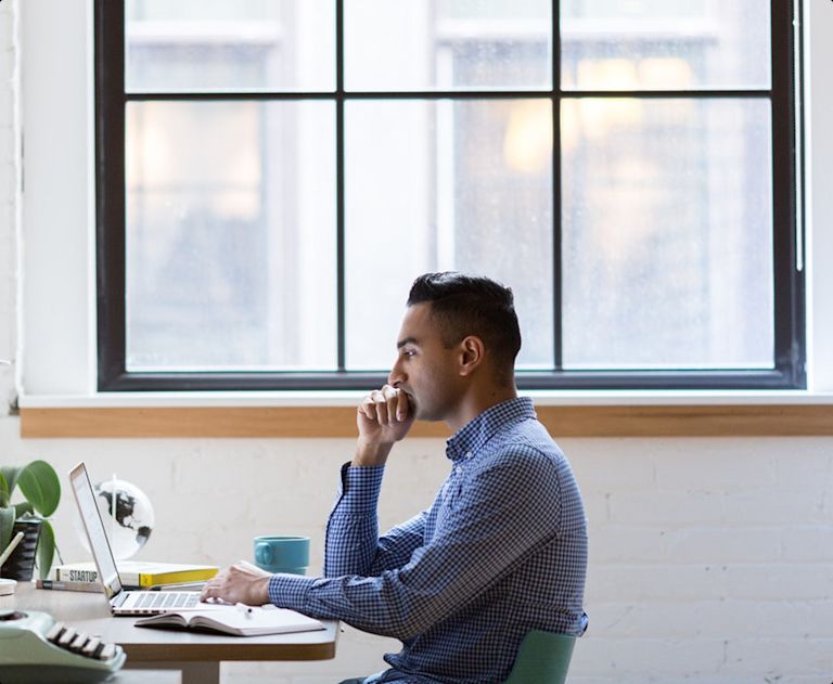 Man sitting at an office desk next to a large window, staring intently at his laptop screen.