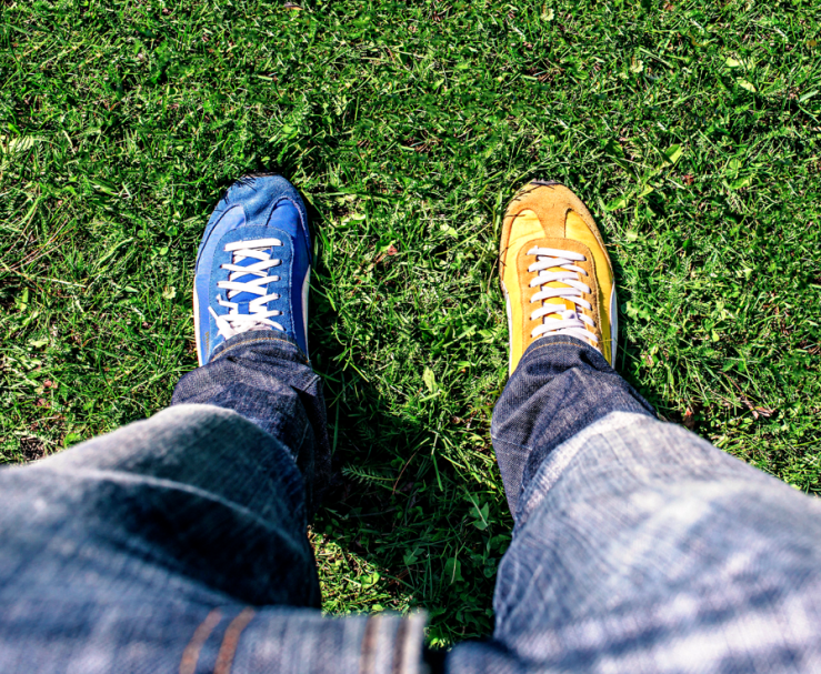 Person stands on the grass, wearing dark blue jeans with one blue and one yellow shoe.