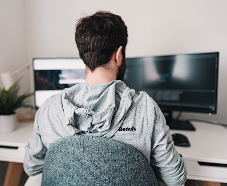 Bearded developer in grey hoodie works at a white desk with two computer monitors.