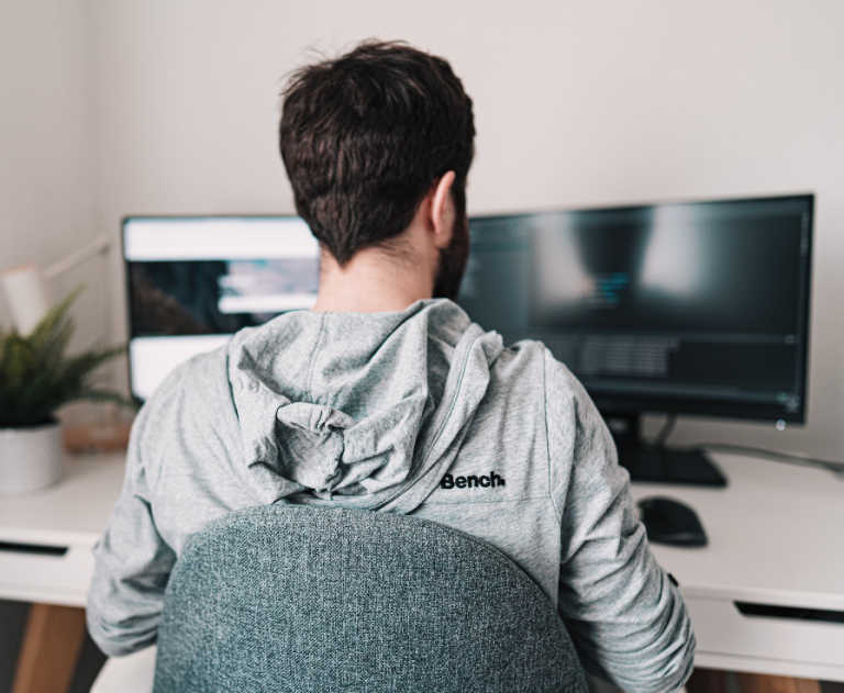 Bearded developer in grey hoodie works at a white desk with two computer monitors.