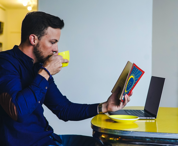Man sips a hot beverage from a yellow teacup, while sitting in front of an open laptop on a yellow table and reading a book.