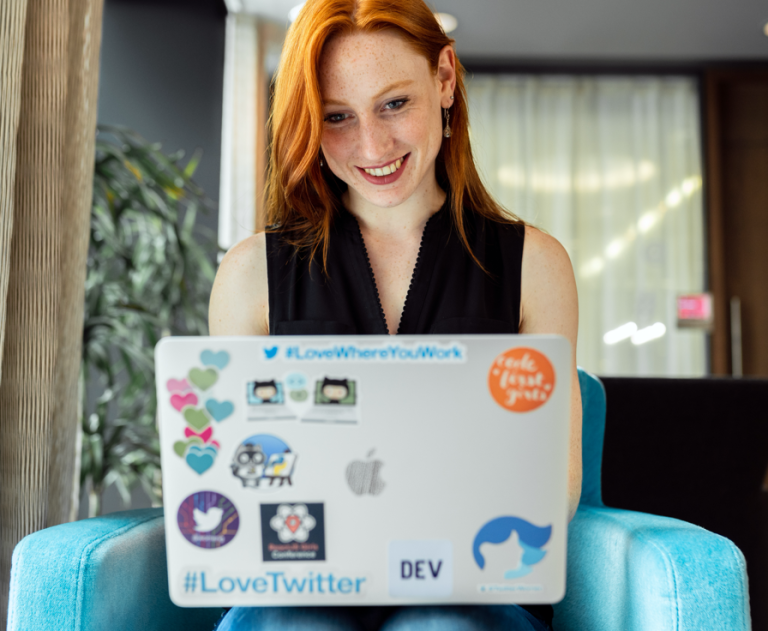 Woman with red hair, black top, and blue jeans sits on a blue armchair and looks at her laptop screen. The back of the screen is covered in stickers including the hashtag #LoveTwitter.