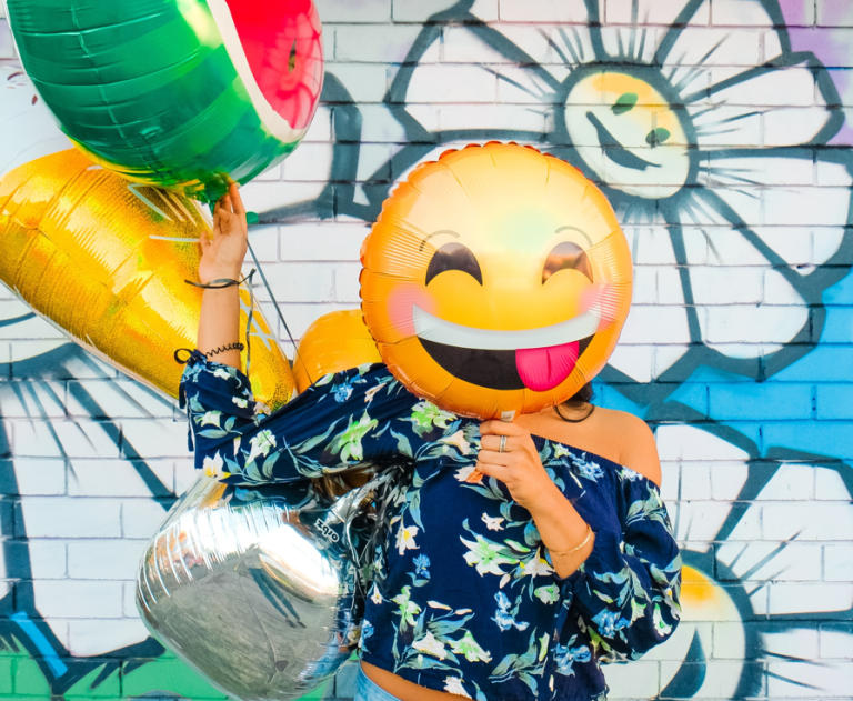 Woman stands in front of a colorful painted brick wall, holding balloons shaped like emoji, with a cheeky-faced balloon covering her face.