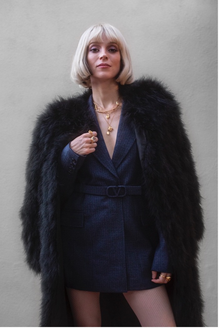 Wearing her blonde wig, St. Vincent stands in front of a Wall and wears a fluffy coat over a suit dress.