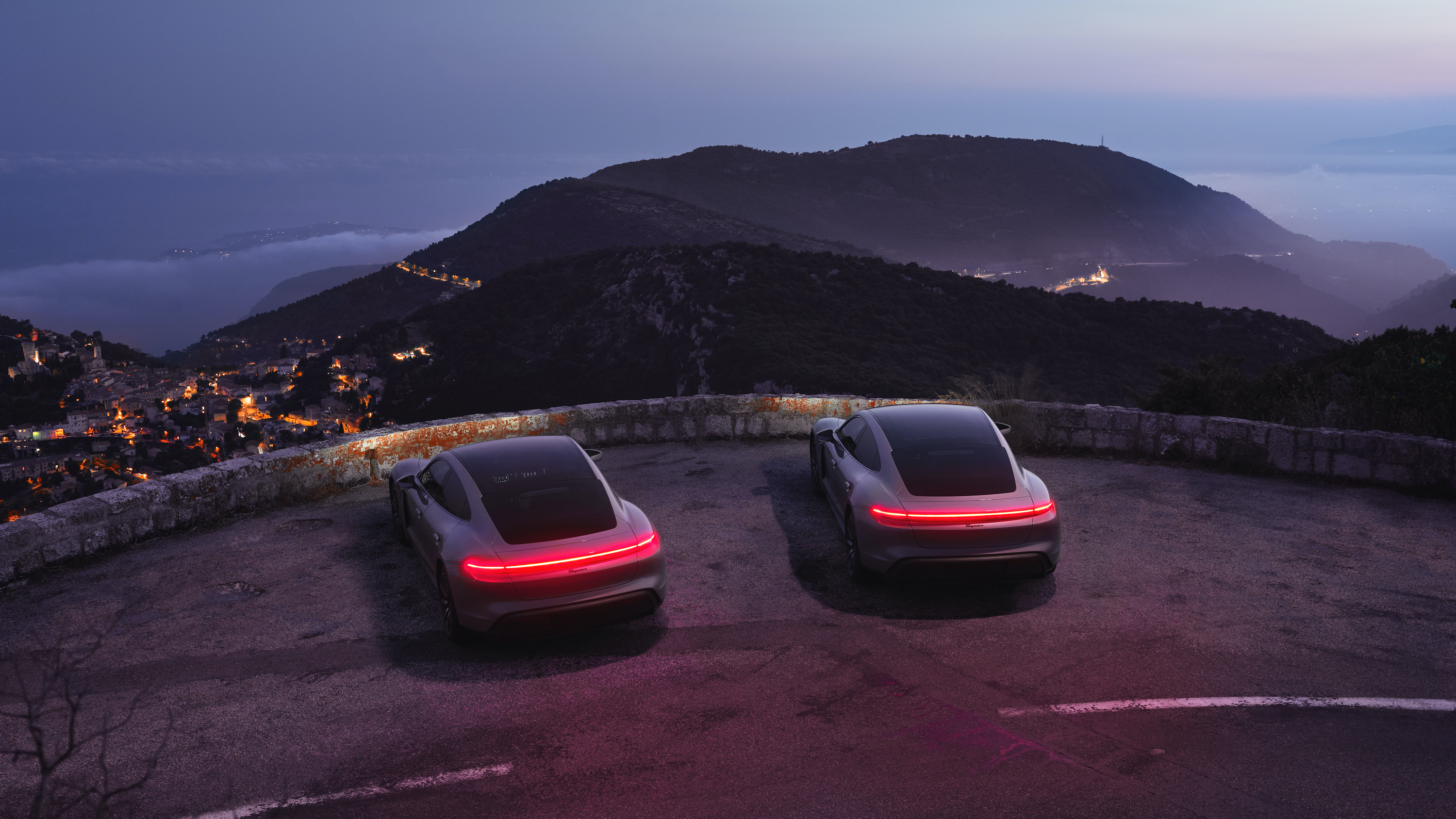 Two Porsche Taycan cars overlook a town at night