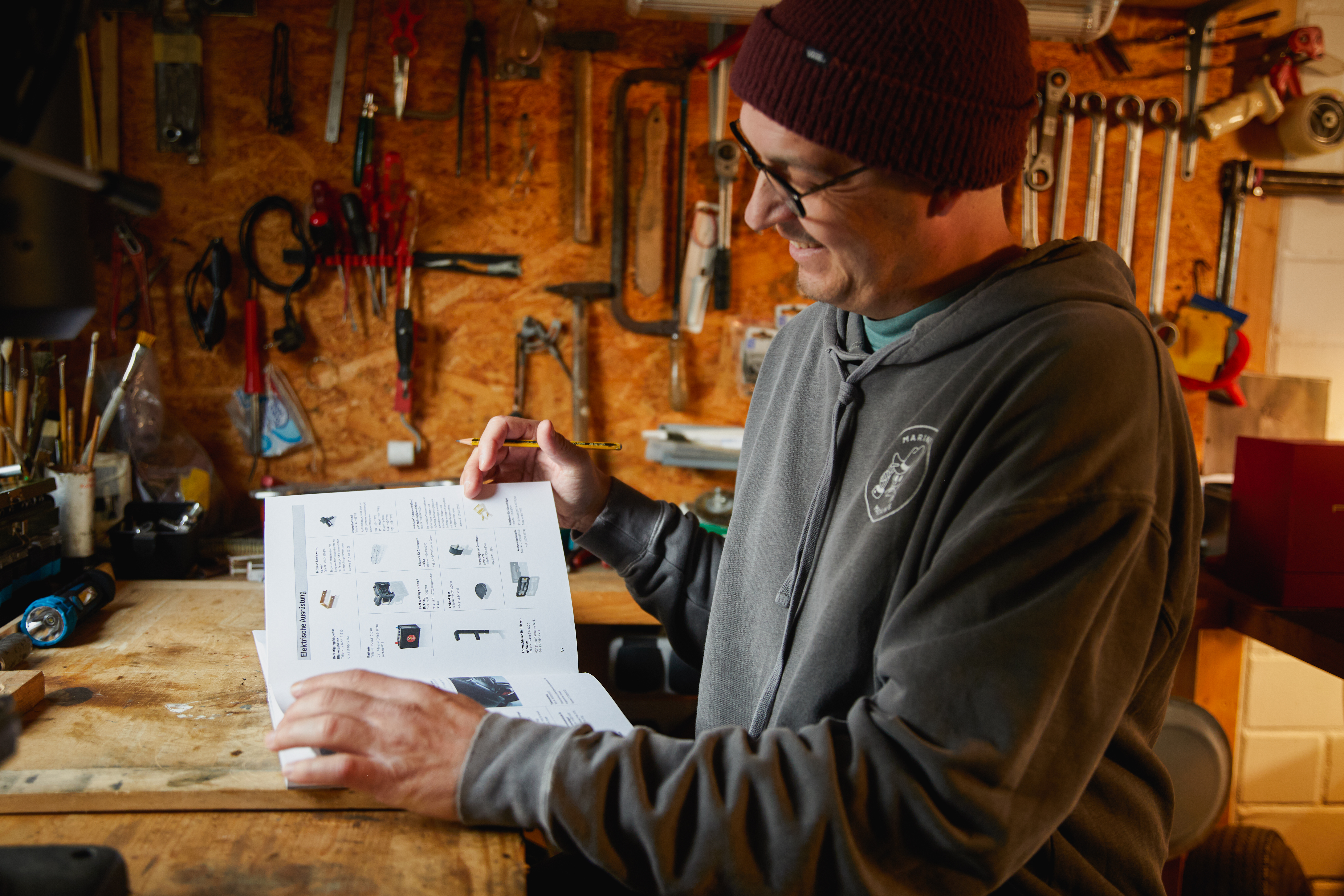 Man in workshop reads magazine, tools hanging on wall behind