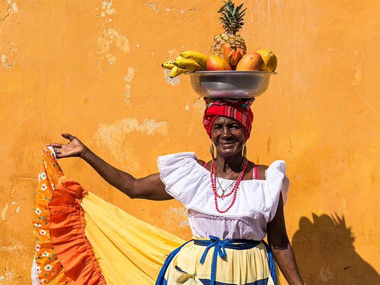Woman carries fruit on head in front of orange wall