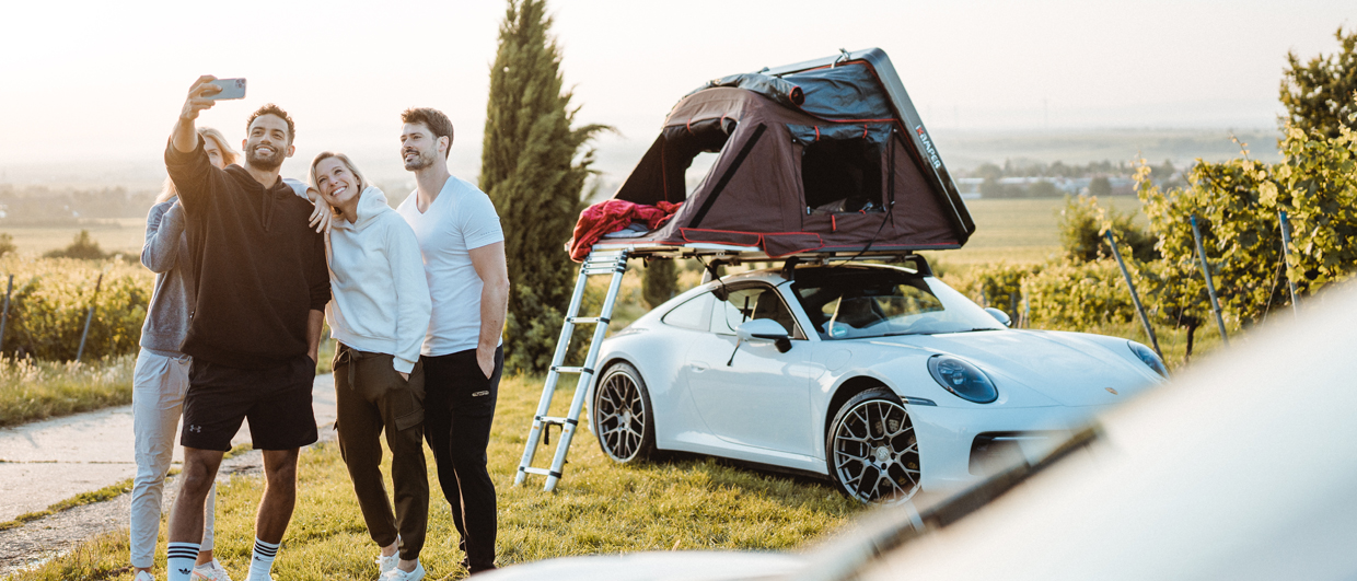 Four people take selfie in front of roof tent-topped Porsche