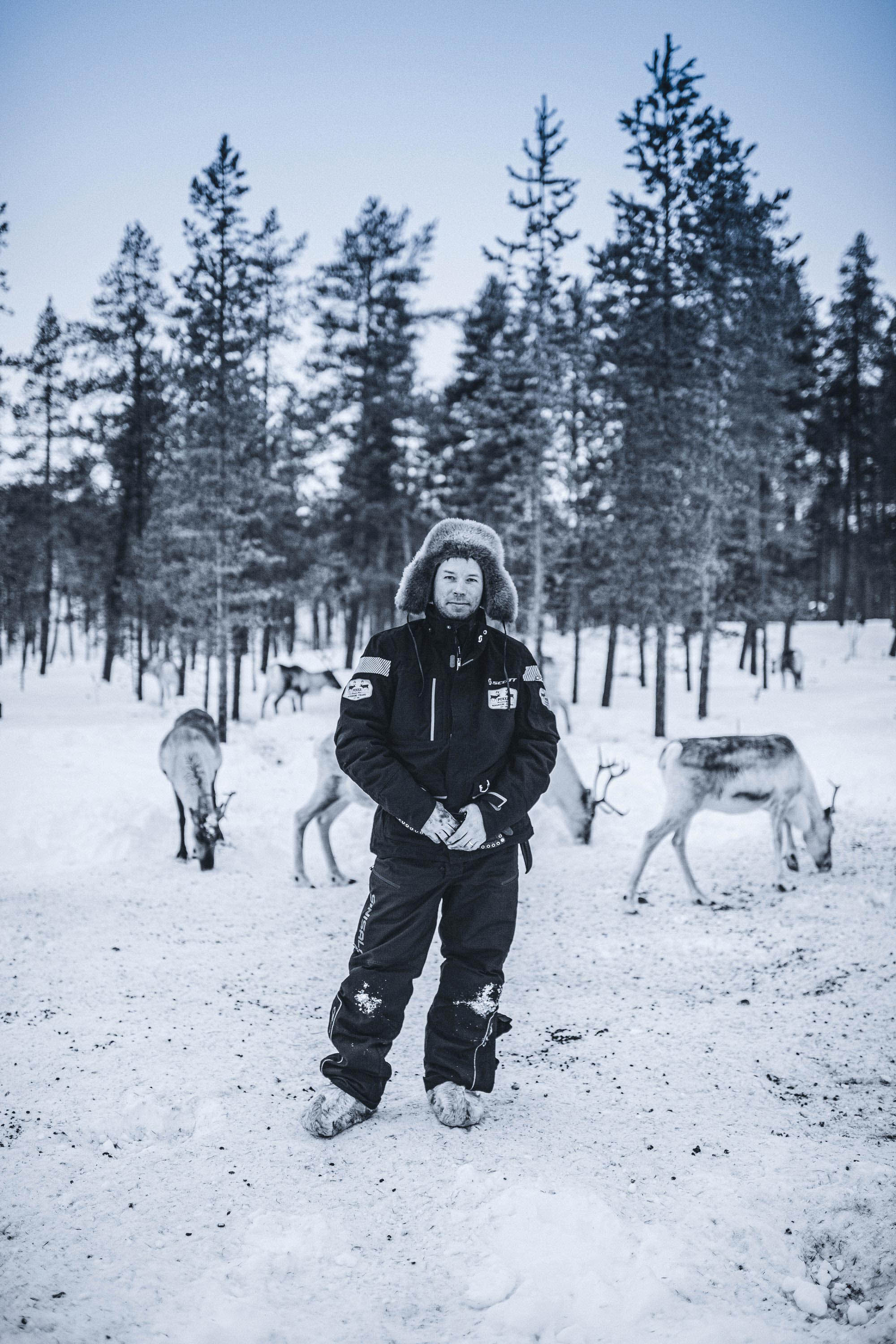 Herdsman in snowsuit, warm fur hat and boots with reindeer behind him
