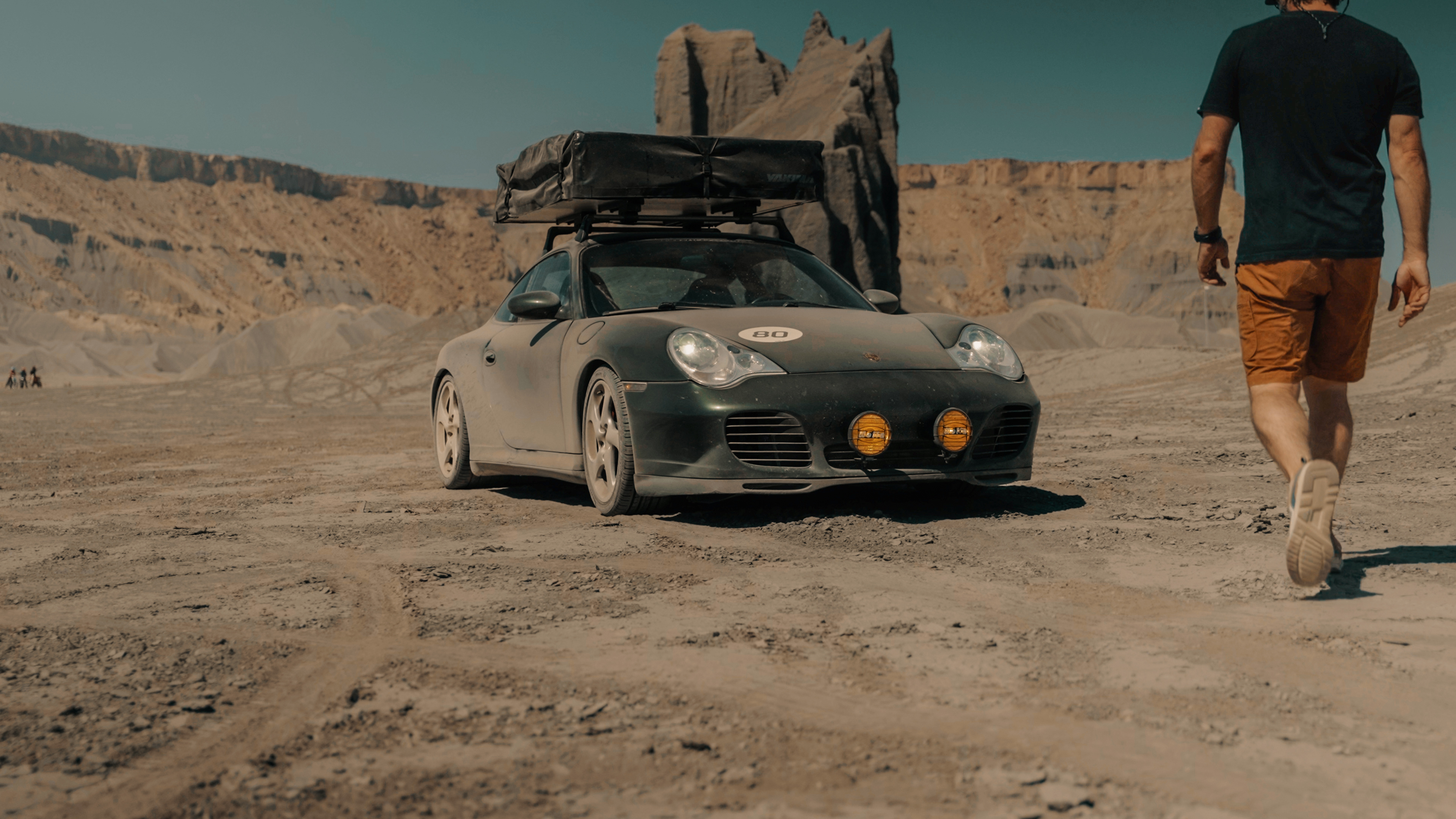 Man walks towards a sandy Porsche in rocky desert