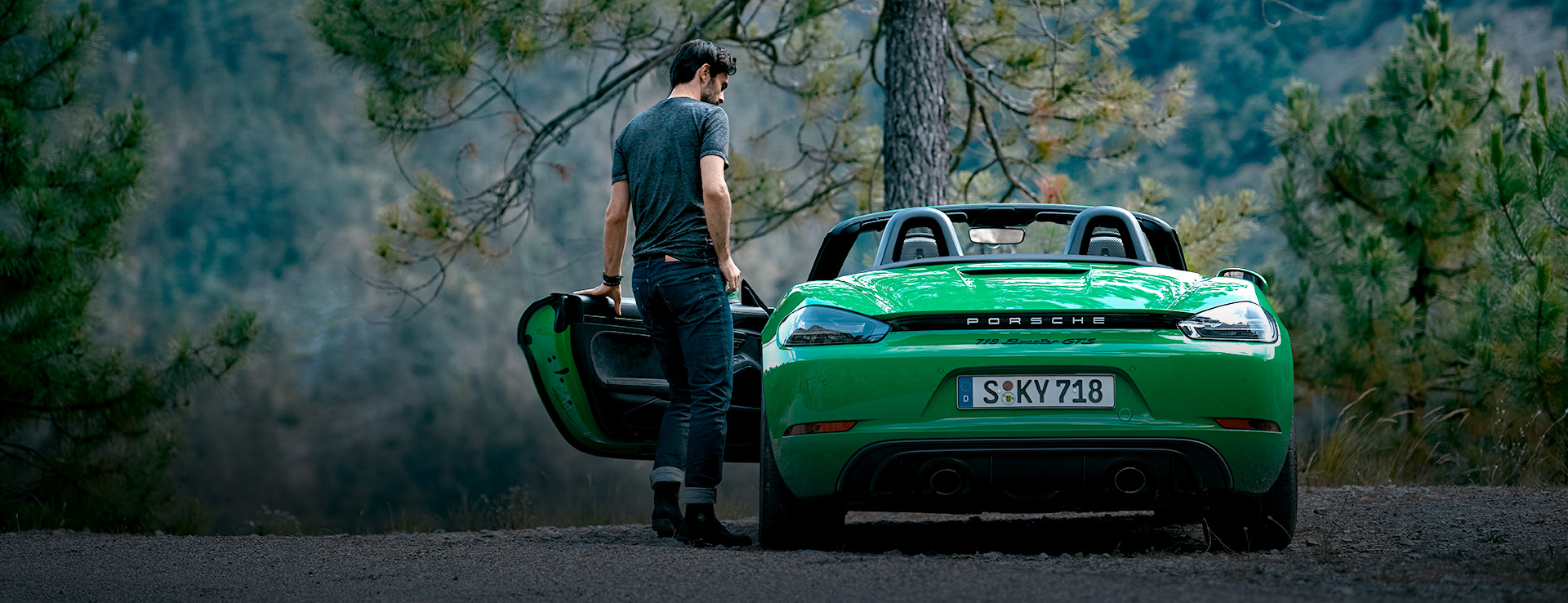 Man entering green 718 Boxster GTS 4.0, pine trees behind