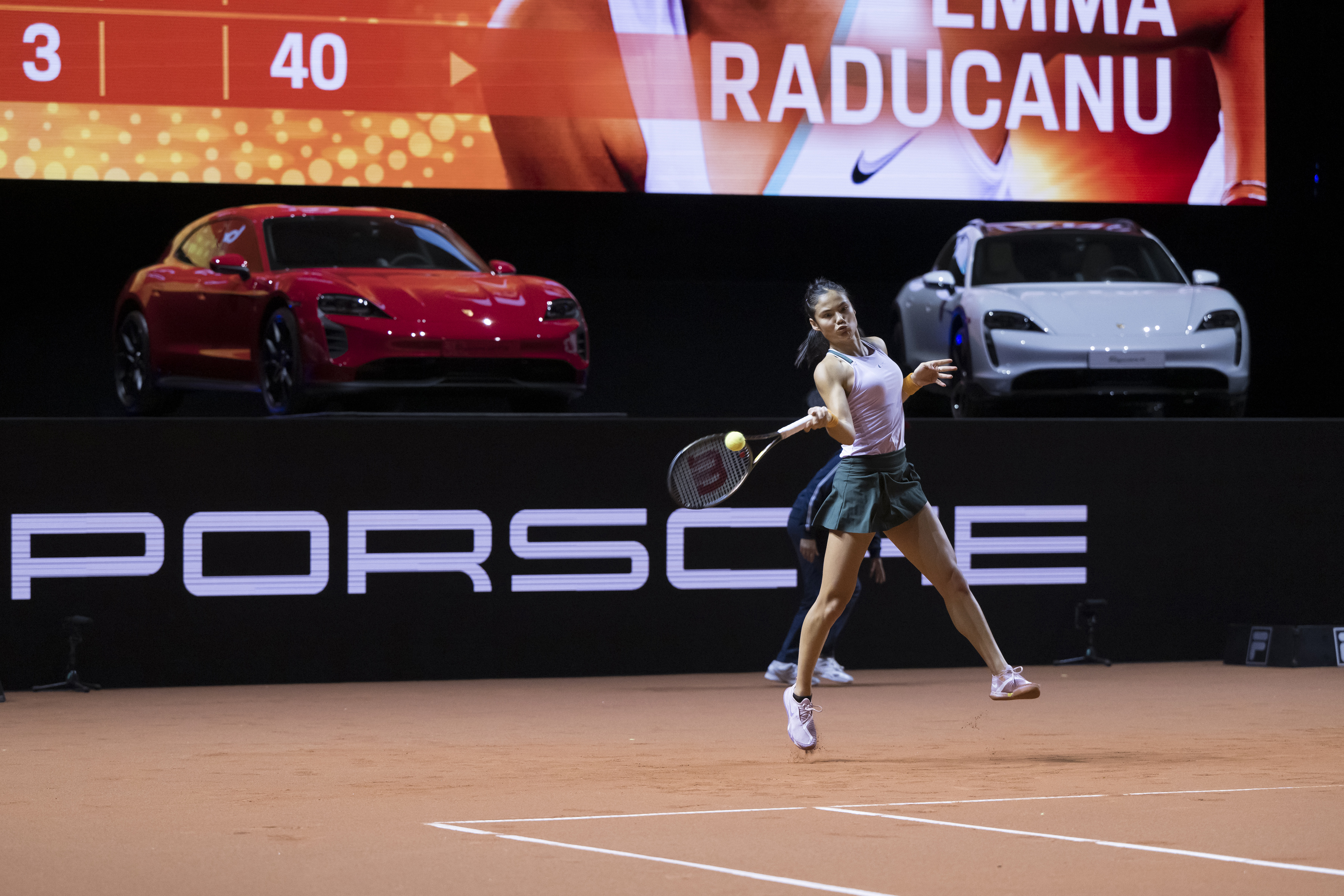 Woman mid-swing on clay tennis court. Porsche Taycans behind.