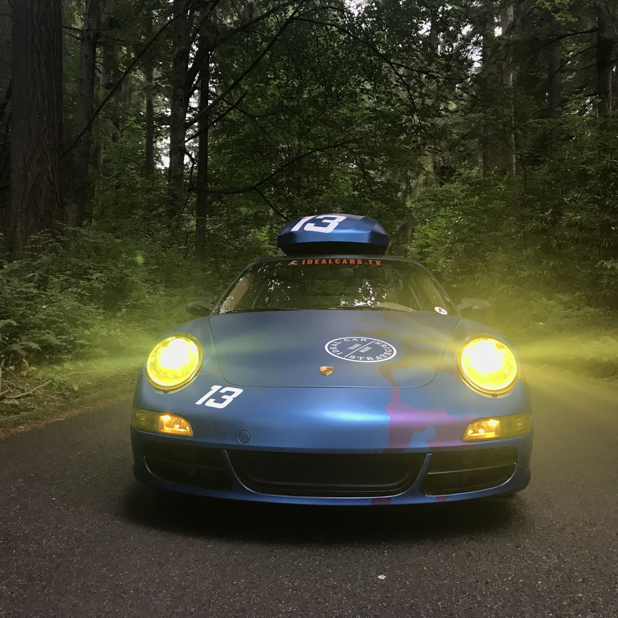 Blue Porsche 911 in a forest at twilight, headlights on
