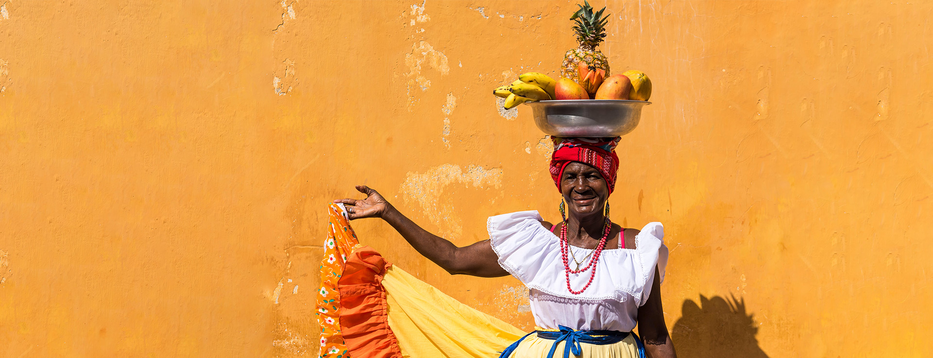 Woman carries fruit on head in front of orange wall