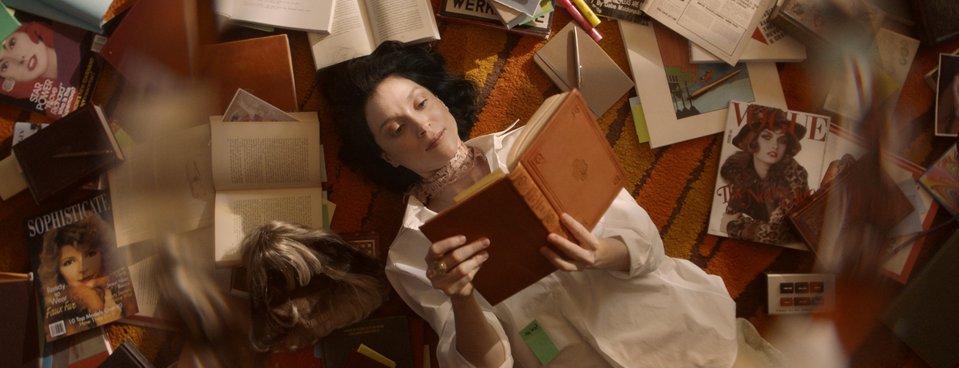 St. Vincent lies on a brightly-carpeted floor in a cozy space. She is surrounded by books and reading a novel.