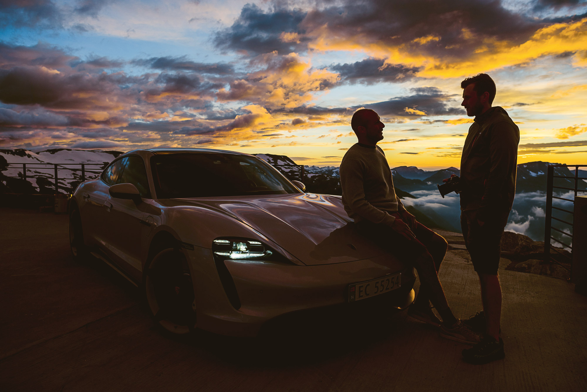 Two men talk, with Porsche Taycan in background, at sunset