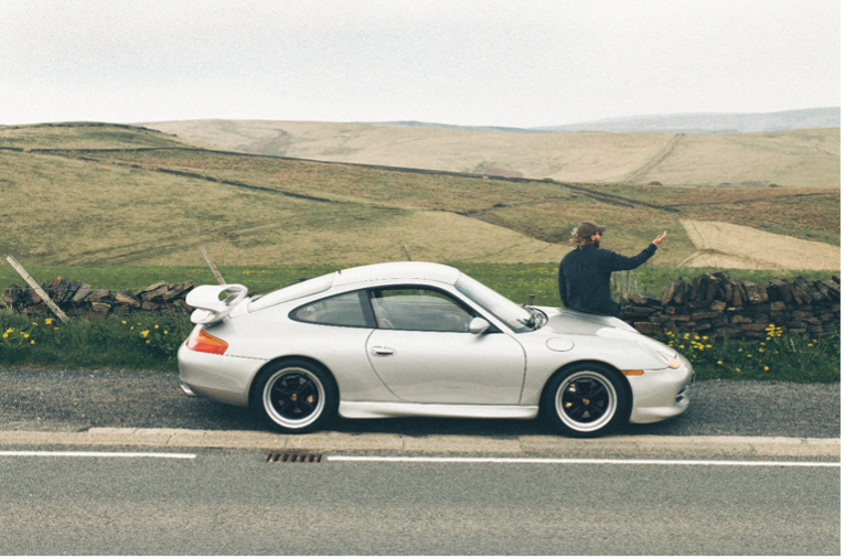 Man sits on bonnet of Porsche 911 (996) overlooking countryside