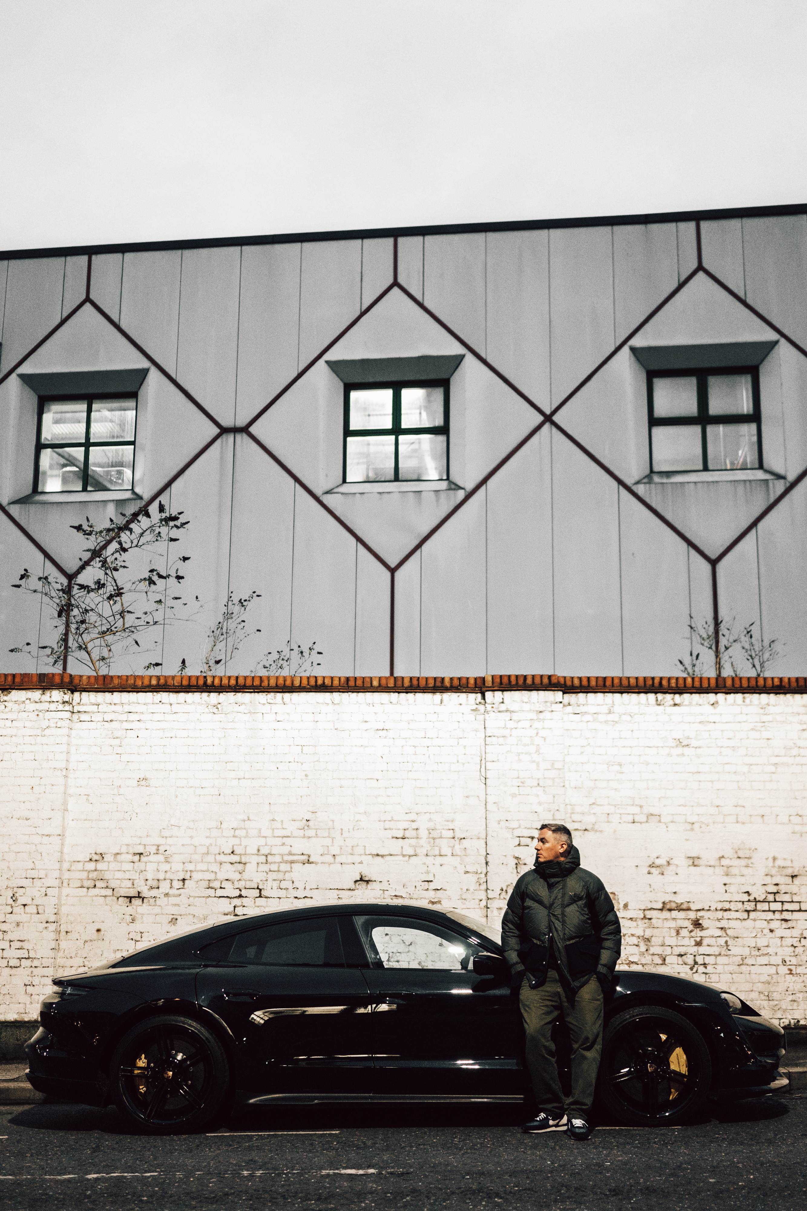 Benji B in padded jacket leaning against black Porsche Taycan