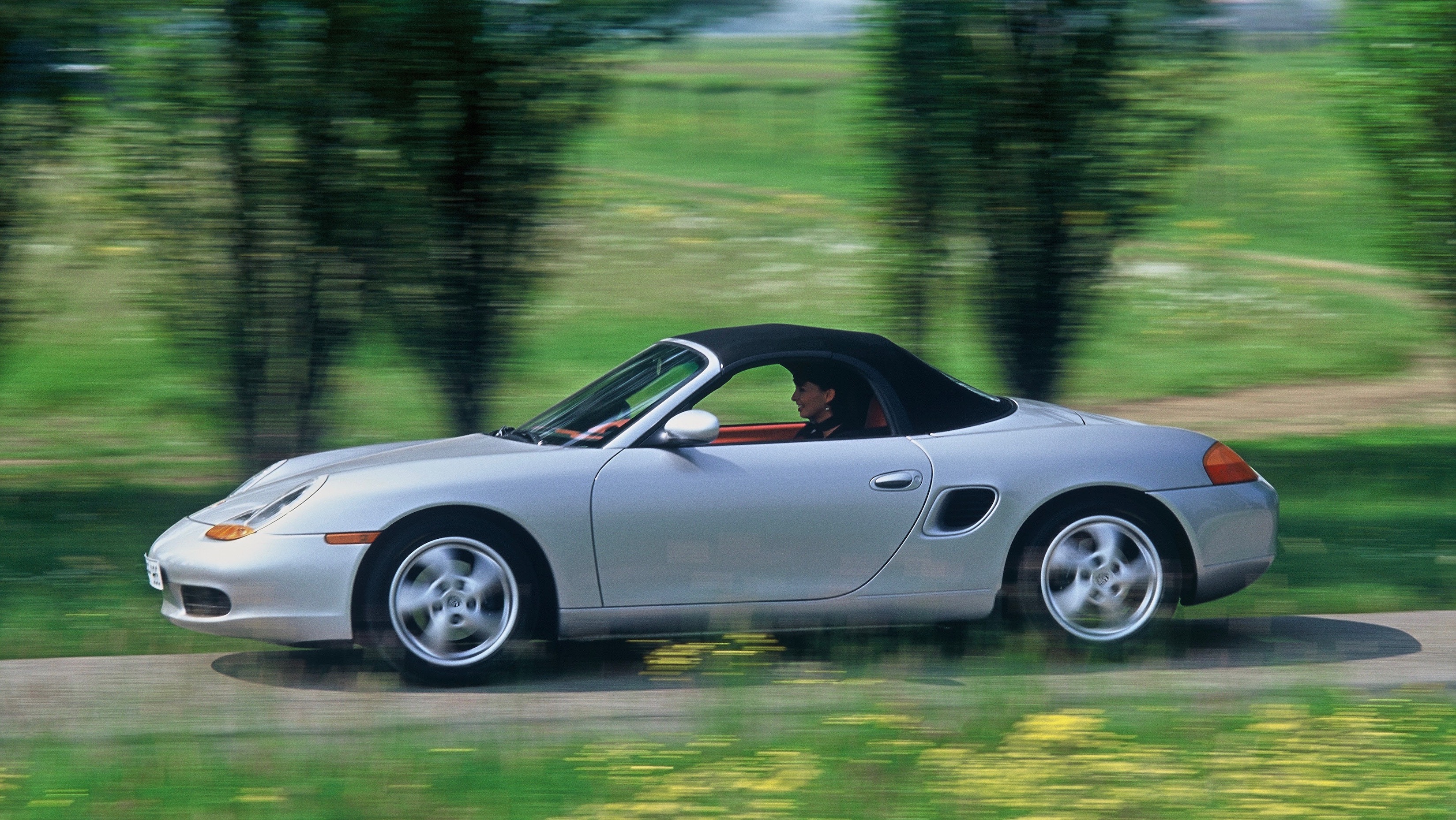 Mk1 Porsche Boxster with roof up driving along tree-lined road