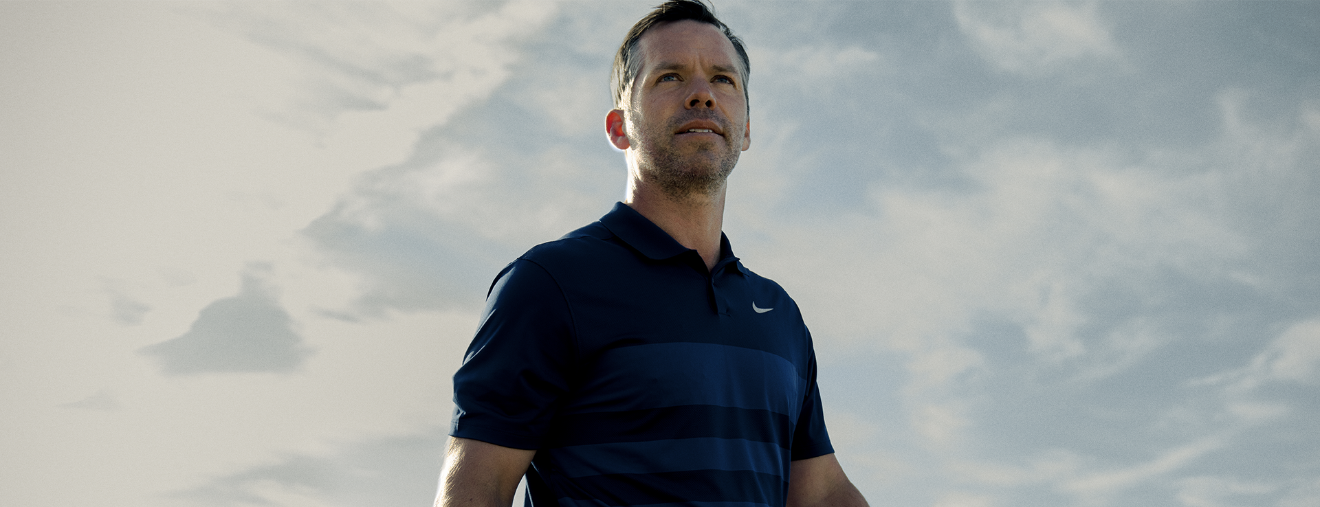 Paul Casey is standing in front of clouds while he looks up into the sky. We see him frontal from a low angle.