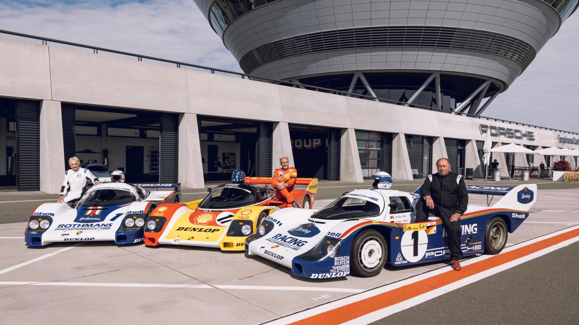 Three racecar drivers standing beside three Group C Porsche cars