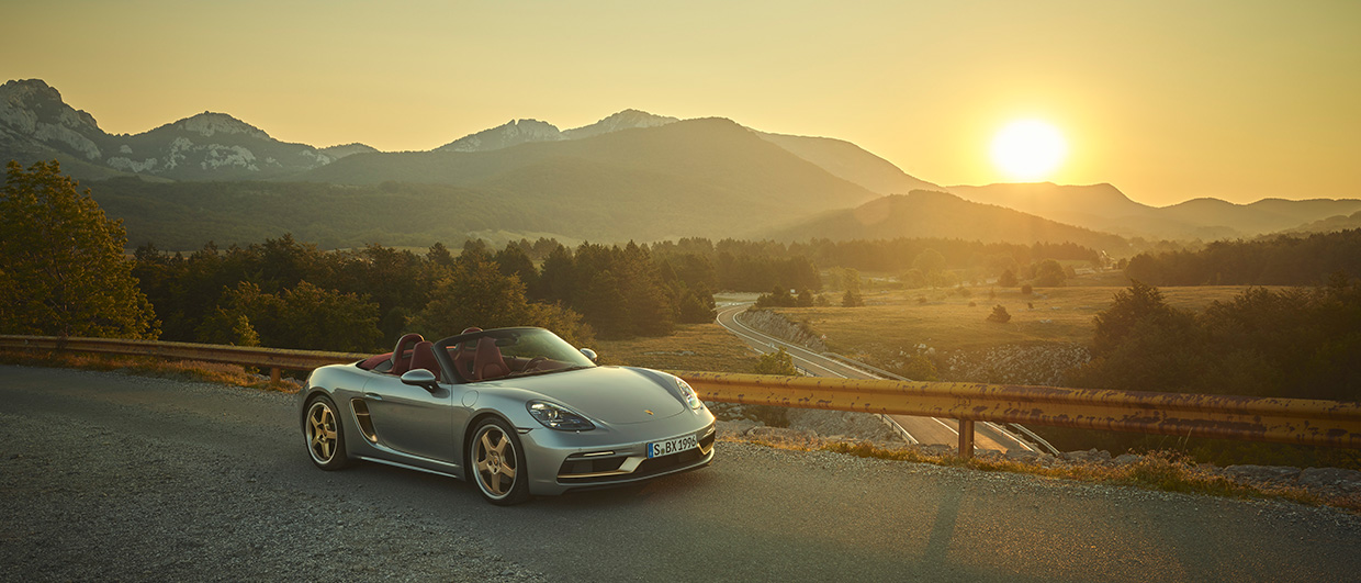 Silver Boxster 25 years lit by setting sun, mountains behind