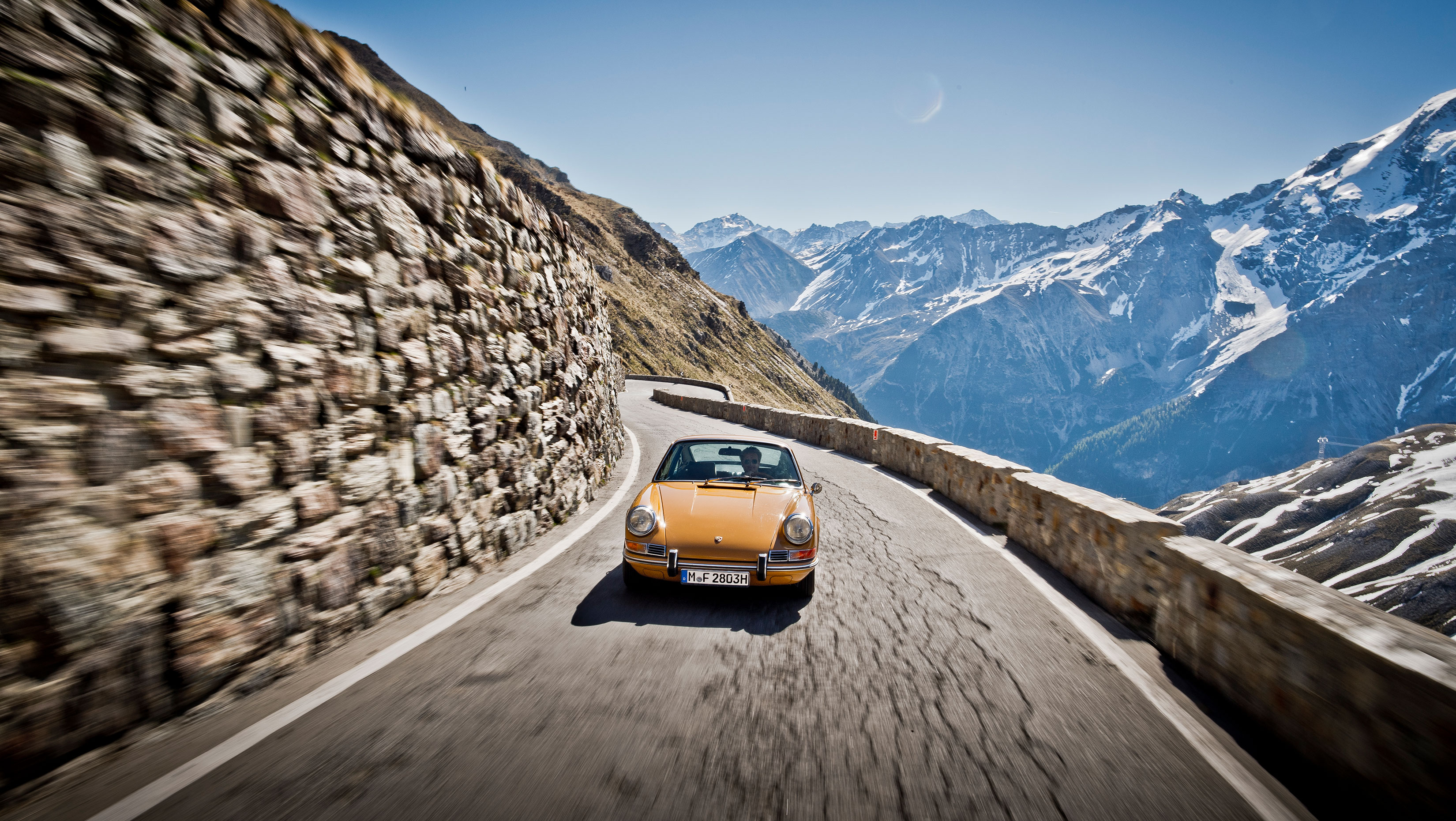 Orange Porsche 911 driving up a wall-lined mountain pass