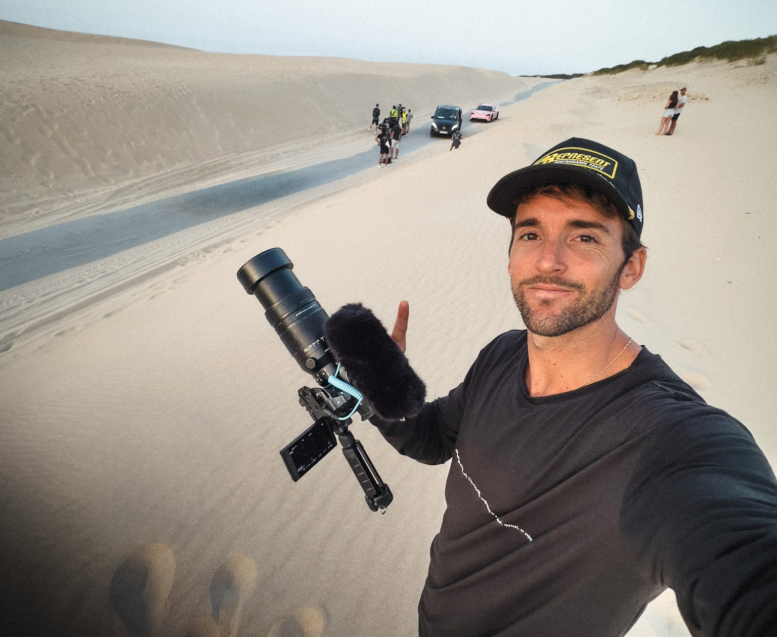 Man holding camera, people, cars and sand dunes behind him
