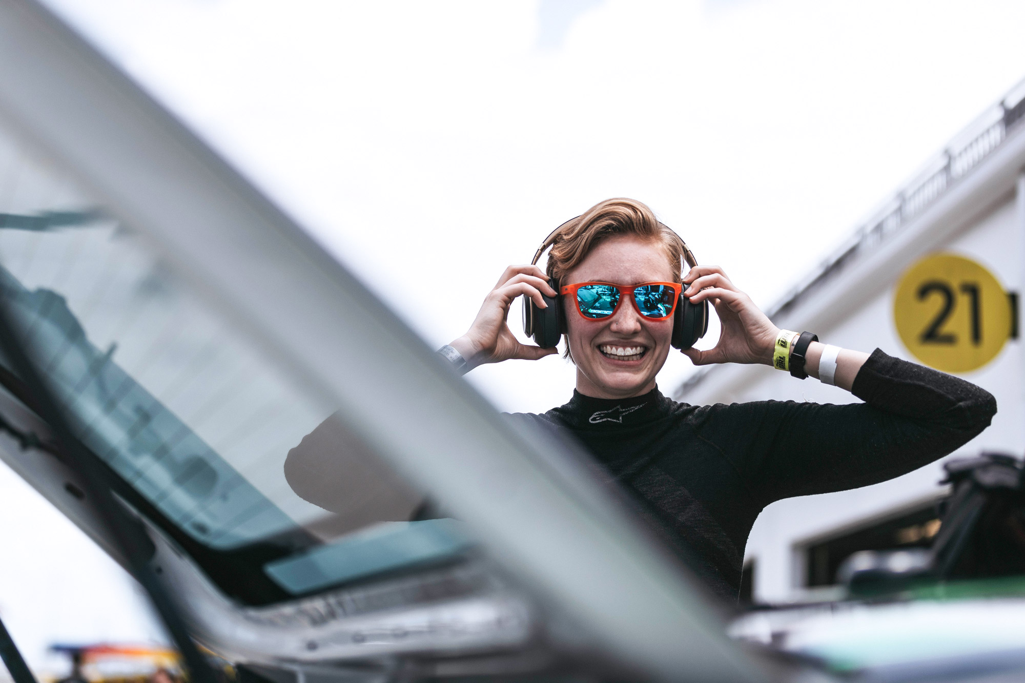 Woman racecar driver, wearing sunglasses, puts headphones on at racetrack