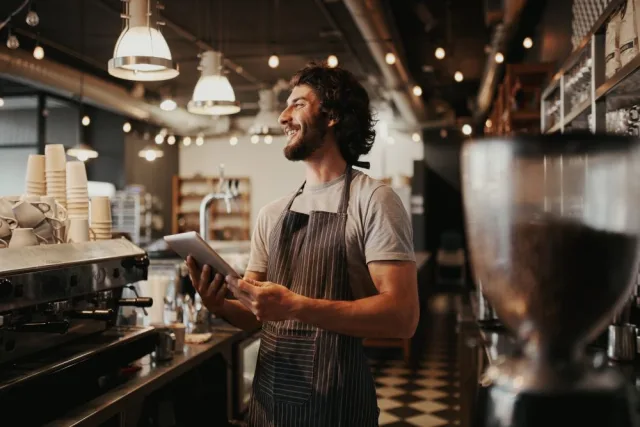 Guy looking away from the camera, standing behind the coffee machine in a coffee shop, smiling and holding an iPad