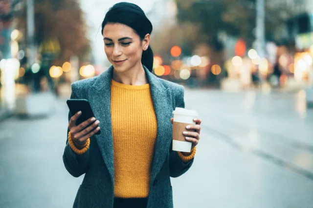 Woman walking on city street texting and holding a coffee