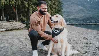 Man kneeling beside Golden Retriever wearing GO! SOLUTIONS bandana on a sandy beach