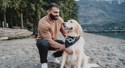 Man kneeling beside Golden Retriever wearing GO! SOLUTIONS bandana on a sandy beach