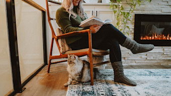 Woman seated reading a book by the fireplace with long haired cat
