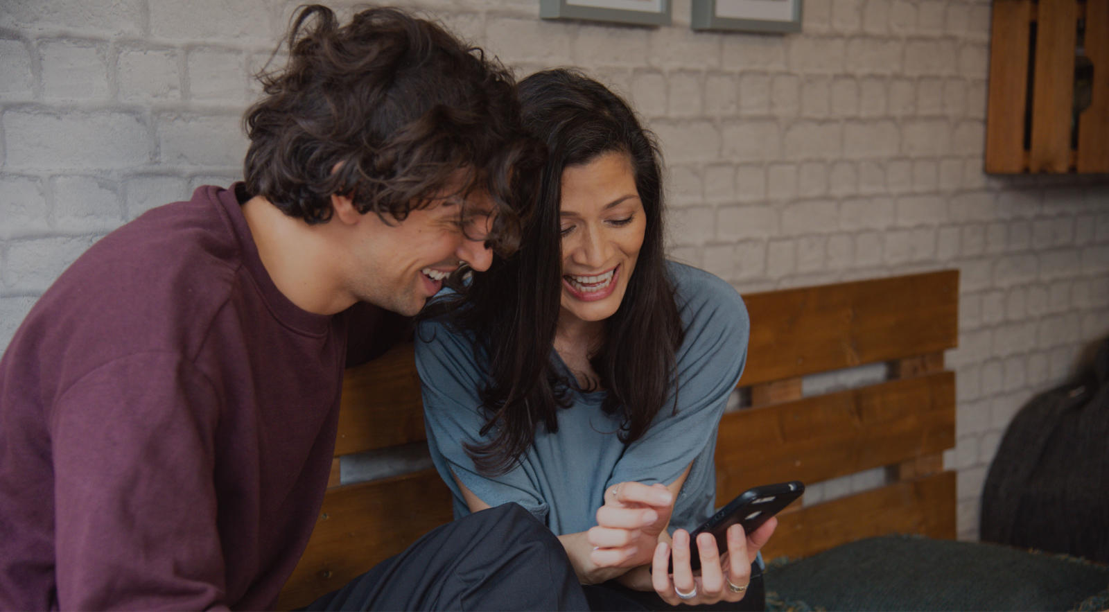 Man and lady sat down, both looking at a mobile phone together and smiling