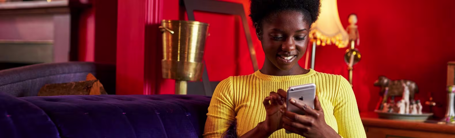 Una mujer africana joven en un sillón morado con un fondo rojo enviando un SMS en el celular.