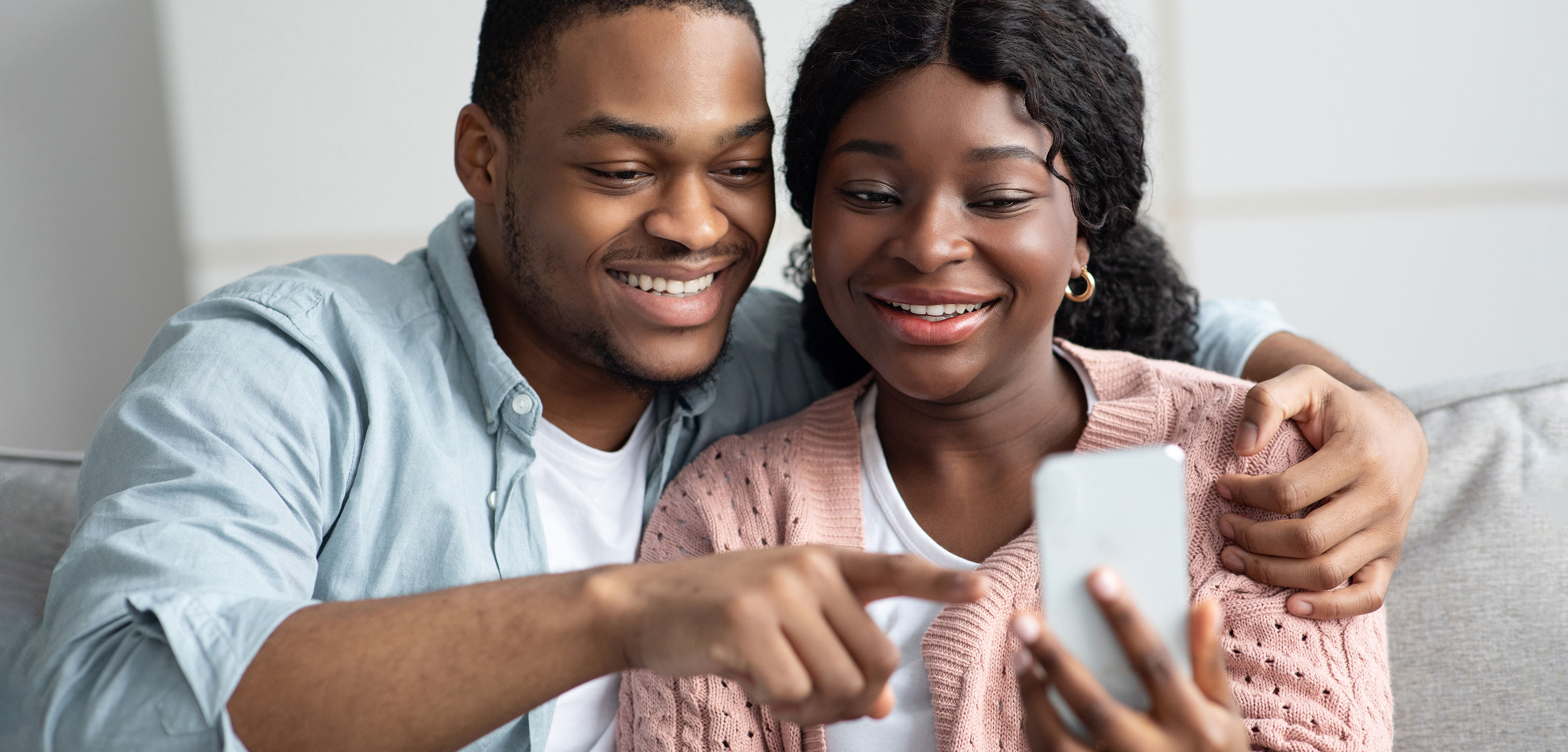 A man sat on the sofa with his arms around a lady, both are looking at a phone