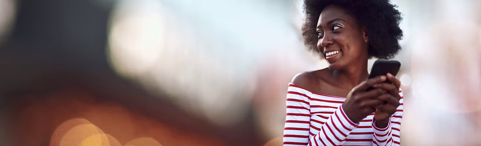 An image of a lady to the far right, looking over her shoulder and smiling whilst holding her phone