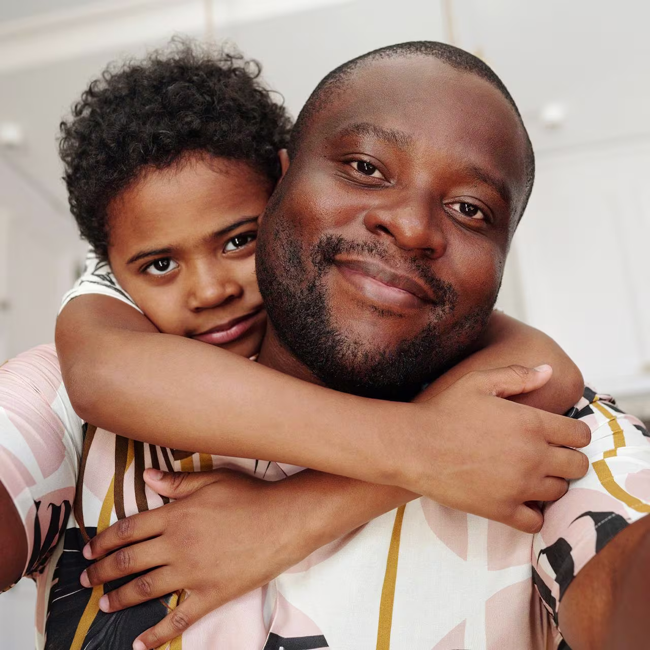 An African man smiling as his son hugs him from the back 