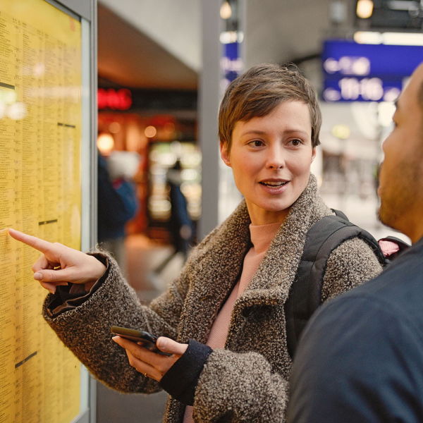 Das Foto zeigt zwei Reisende auf Bahnsteig an der Fahrplanauskunft. Links steht eine Frau mit kurzen Haaren. Sie zeigt mit ihrem Finger auf den Fahrplan und schaut einen Mann an, der rechts neben ihr steht. 
Copyright: Deutsche Bahn AG/ Max Lautenschläger