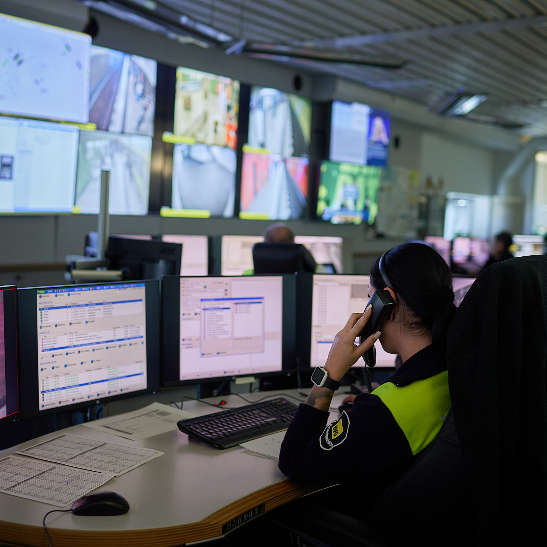 Man sieht eine Frau, die vor vielen Bildschirmen sitzt und dabei telefoniert.