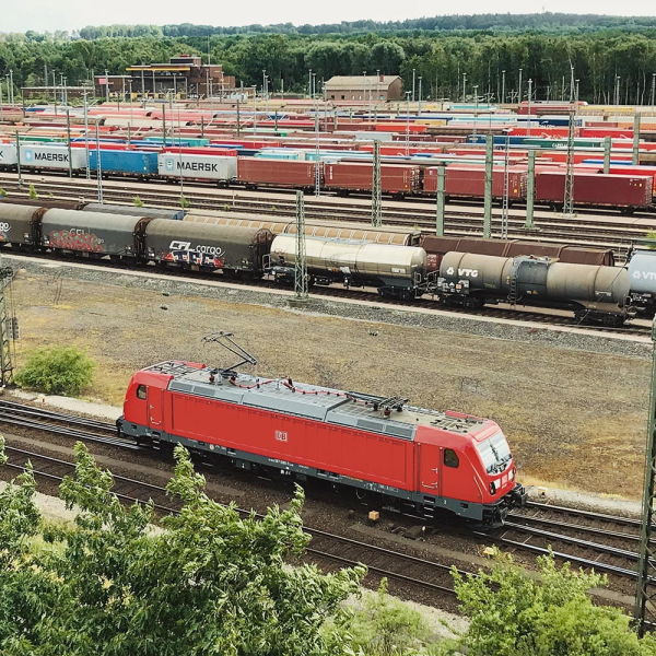 Das Bild zeigt einen roten Triebwagen auf einem Gleis im Rangierbahnhof Maschen und die DB Cargo Ellok Baureihe 187 rollt zum nächsten Einsatz. Im Hintergrund stehen viele Waggons des Güterverkehrs.
Copyright: Deutsche Bahn AG/ Volker Emersleben